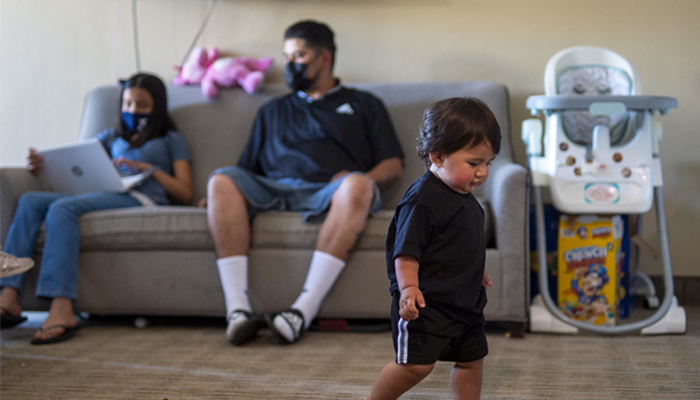 Young child walking in a living room with parents seated in the background.